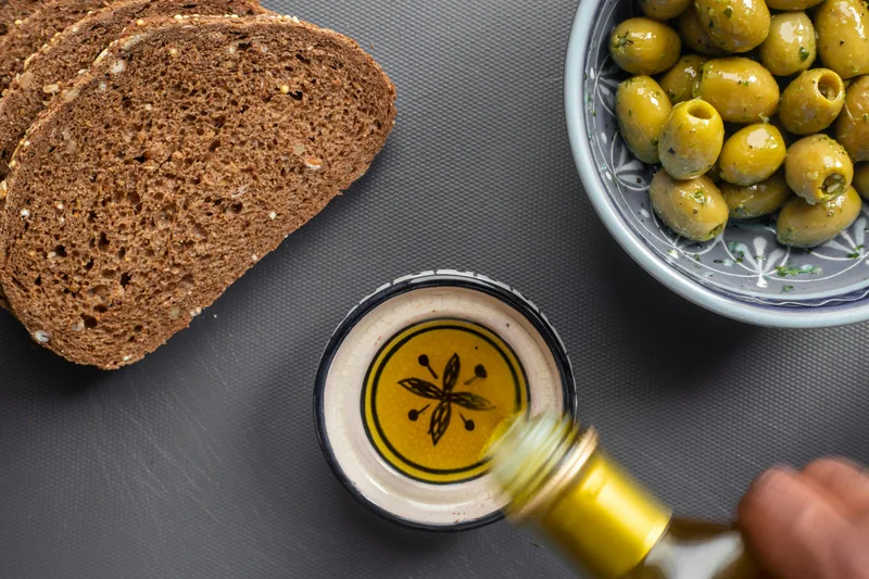 Olive oil poured into a bowl beside bread and green olives
